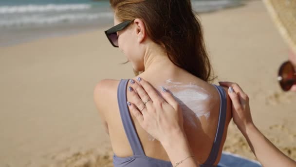 在沙滩上给女人涂防晒霜的女孩 微笑的女士准备在海滨晒日光浴 把防晒霜涂在皮肤上 度假概念 防晒护肤 — 图库视频影像
