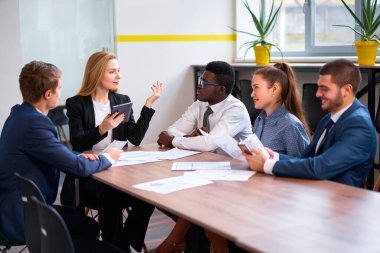 Genç kadın toplantıyı yönetiyor, takım strateji konusunda işbirliği yapıyor. Çok ırklı profesyoneller projeyi modern ofiste tartışırlar. Tabletli meslektaşlar, kağıtlar takım çalışması, kurumsal tartışma.