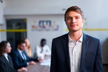 Şirket toplantısında takım elbiseli lider. Kendine güvenen genç işadamı ofiste duruyor, ekip arkadaki masada işbirliği yapıyor. Profesyonel grup, modern çalışma alanında tartışma ve takım çalışması yapıyor..