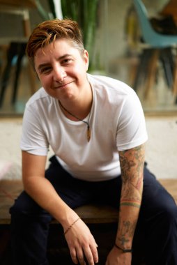 Gülümseyen, kapsayıcı iş yeri, cinsiyet çeşitliliği kutlandı. Kendine güvenen transseksüel profesyonel şehir ofisinde oturuyor. Dövmeli, gündelik tarzda, içten içe doğal. Başarı, modern iş atmosferi.