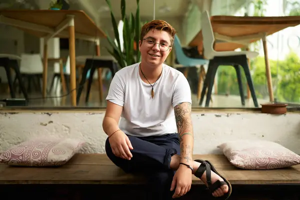 Stock image Tattooed gender-fluid person displays comfort, inclusivity with airy office space. Smiling Gen Z transgender office worker in smart casual clothes sits on bench, modern workplace environment.