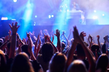 Hayranlar canlı performansı, gece enerjisini, yaz etkinliğini severler. Konsere katılanlar, açık hava müzik festivalinde ritim içinde el kaldırıyor, renkli sahne ışıkları heyecanlı seyircileri aydınlatıyor.