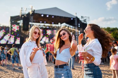 Güneş gözlüklü gülümseyen bayan arkadaşlar soğuk içecekler tutarlar, güneşli bir günde sahnenin yanında dans ederler. Bir grup kız kumsalda yaz müzik festivalinin tadını çıkarıyor. Parti, eğlence, deniz kıyısındaki gençlik yaşam tarzı.
