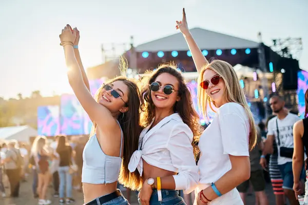Kadın arkadaşlar konserden, kalabalıkla çevrili eğlenceli zamanlardan, gün batımında okyanus arkaplanından hoşlanırlar. Üç neşeli kız güneşli yaz plaj müzik festivalinde dans ediyor, kollarını kaldırıp kutlama yapıyorlar..