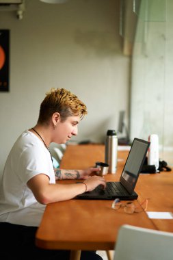 Yaratıcı ofis ortamında gündelik profesyoneller dijital görevlere girişiyor, içecekten hoşlanıyor, çevik iş kültürünü yansıtıyor. Transseksüel serbest çalışan biri laptopta çalışır, elektrikli süpürge bardağından çay yudumlar..