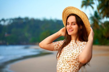 Rahat turistler deniz kenarında sıcak bir akşamın tadını çıkarırlar. Gün batımında tropik plajda hasır şapkalı gülümseyen genç bir kadın. Doğada eğlence, yaz seyahati, tatil havası. Palmiye ağaçları ile canlı okyanus arkaplanı.