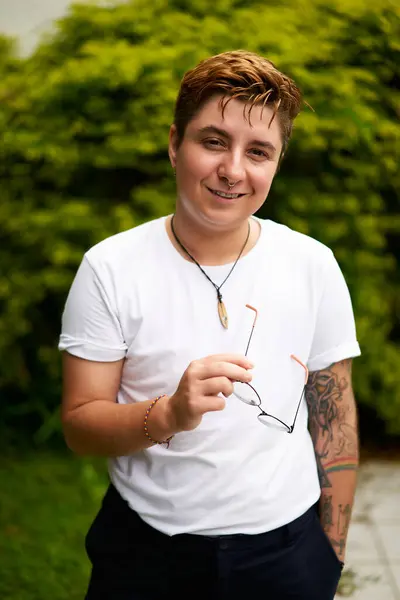 Smiling transgender man outdoors in casual attire, confidence evident in stance with glasses in hand, tattoo visible on arm, plants backdrop suggesting fresh start, inclusivity gender identity pride.
