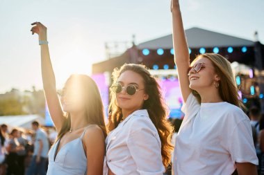 Kadın arkadaşlar sahildeki yaz müzik etkinliklerinden hoşlanırlar, altın saat ışığı, neşeli kalabalık titreşimleri. Gün batımında güneş gözlüklü kızlar, kollar havada, kumlu sahilde dans eden festival sahnesi geçmişi olan kızlar..
