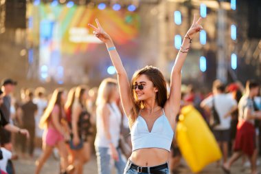 Kollar havada, barış işareti, sahne ışıklarıyla kalabalık içinde dans. Mutlu genç kadın plajda yaz müzik festivalinin tadını çıkarıyor. Günlük moda, güneş gözlüğü, arkadaşlarla gün batımı konser partisinde eğlence..