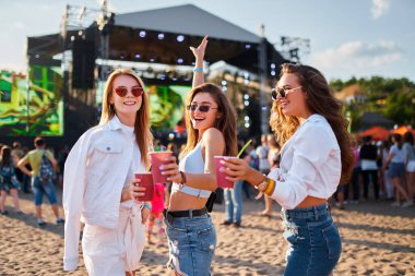 Yaz kıyafetleri içinde gülen katılımcılar, okyanus kenarında keyifli bir açık hava festivali havası. Kadınlar, güneşli sahil müzik etkinliklerinde kokteyllerle kadeh kaldırır. Gösterinin, arkadaşlığın ve eğlencenin tadını çıkarırlar..