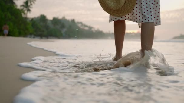 海のサーフウォーターエッジで裸足で日没する女性の足 泡立った水を散らす 若い女性の足は夏の夕方に海岸沿いにゆっくりと歩いています 海辺で休暇を楽しむドレスの女性 — ストック動画