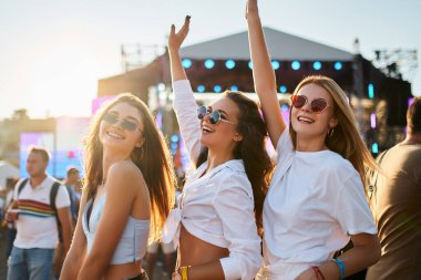 Arkadaşlar dans eder, yaz etkinliğini kutlar, kollarını kaldırır. Plaj müzik festivalindeki bir grup neşeli kız. Gülümseyen yüzlerde parlak güneş ışığı. Gençlik sevinci, canlı konser eğlencesi. Sıradan moda, arkadaşlık anları..