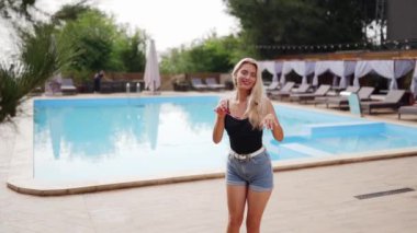 Sarı saçlı kadın kırmızı güneş gözlüğü, şort ve yüzme havuzunda güneş pansiyonları ve çam ağaçlarıyla fotoğraf çekmek için dans ediyor. Tatillerde poz vermek ve flört etmek.