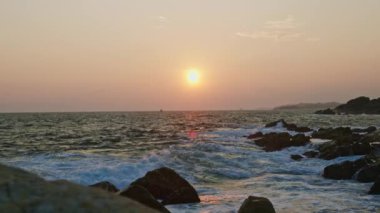 Titrek gökyüzü alacakaranlıkta denizle buluşur, suyu yansıtır. Fırtınalı okyanus dalgaları üzerinde gün batımı ufku, ön planda kayalar. Sakin ama güçlü bir doğa sahnesi. İnsanlar, vahşi sahilin ebedi cazibesi. Yavaşla