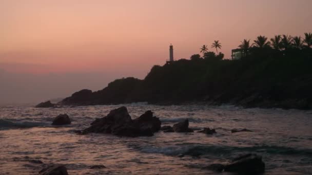 黄昏时海浪冲击着岩石海岸 形成了平静的海上自然风光 灯塔耸立在山上 夕阳西下 天空漆成粉红色 棉花糖的色调营造了宁静的氛围 — 图库视频影像