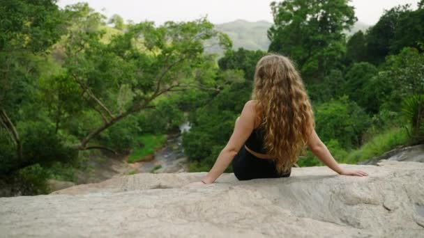 女性は静かな瞬間 ソロ旅行の冒険 瞑想的なリトリート 森の水をカスケードするエコツーリズムを楽しんでいます 長髪の女性は風光明媚な滝の端でリラックスし ジャングルの自然を眺めます スローモ — ストック動画