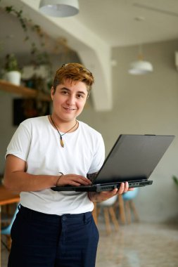Short-haired, smiling FTM pro in casual wear. Confident transgender employee holds laptop in modern office cafe. Inclusive workplace, gender diversity, tech job. clipart
