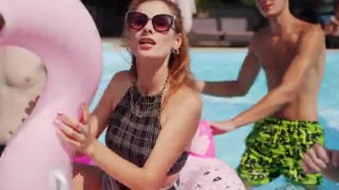 Güneş gözlüklü ve bikinili bir manken yaz havuz partisinde bir grup arkadaşla şişme flamingo tüpünde yüzüyor. Çekici kadın poz veriyor ve turkuaz mavi suda pembe kauçuk halkayla şarkı söylüyor..