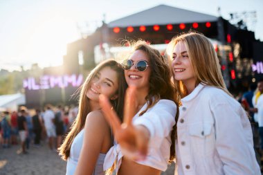 Neşeli bir plaj müzik festivalinde güneşli bir günün keyfini çıkaran, selfie çeken, dans eden ve arka planda sahne alan festivale katılanların arasında arkadaşlığı kutlayan bir grup mutlu genç kadın..