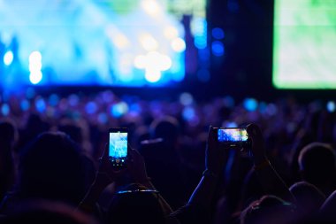 Taraftarlar açık hava festivalinde canlı müzik kaydediyor. Akıllı telefonlar, sahne ışıkları heyecanlı seyircileri aydınlatıyor. Göstericiler sahnede eğleniyor..