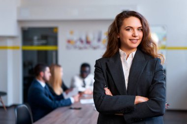 Sarışın patron takım toplantısını yönetir, kadın liderliği somutlaştırır. Kendine güvenen beyaz iş kadını ofiste duruyor, kolları bağlı. Çeşitli personelle modern ofis stratejileri tartışıyor.