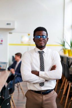 Resmi giyinmiş gülümseyen işadamı, çeşitli takım ortamları arasında liderlik ve profesyonelliği somutlaştırıyor. Kendine güvenen Afrikalı şirket çalışanı, kollarını kavuşturdu, moda ofis çalışma alanında..