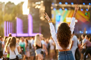 Plaj konserinde kalabalık danslar, gün batımı. Kaldırılmış elleri olan neşeli kadının arka görünüşü yaz müzik festivalinin keyfini çıkarıyor. Açık havada canlı yayında mutlu hayran tezahüratları. Heyecanlı katılımcı kutluyor, titreşimler.