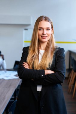 Kollar bağlı, liderlik somut, özgüven. Modern ofiste şık giyinen kendine güvenen sarışın kadın duruyor. Profesyonel şirket ortamı. İş arkadaşları arkada çalışır.