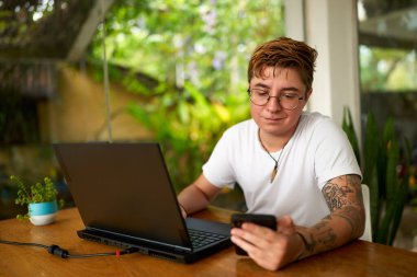Sıradan iş ortamı, iş yerinde cinsiyet çeşitliliği. Dövmeli transseksüel ofis çalışanı akıllı telefonu kontrol ediyor, dikkati laptoptan dağılıyor. Kapalı alan bitkisi, doğal ışık, modern işyerine dahil etme.
