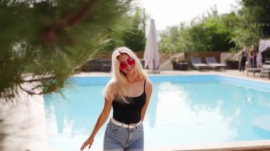 Sarı saçlı kadın kırmızı güneş gözlüğü, şort ve mayo ile fotoshoot için dans ediyor. Plaj havuzunda güneş pansiyonları ve çam ağaçları olan bir mayo. Tatillerde poz vermek ve flört etmek.