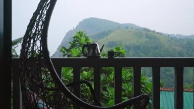 Dolly shot, lüks terası, sallanan sandalyeyi, Ella Rock manzarasını gözler önüne seriyor. Tırabzanda çaydanlık ve bardak, Sri Lanka dağ manzaralı çay demleme. Huzurlu bir sabah, çay severler manzaralı dağlarda keyif alırlar. Yavaşla