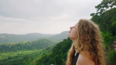 Yeşil orman manzarasının tadını çıkaran bir kadınla huzurlu doğa sahnesi. Kıvırcık saçlı kadın uçurumun kenarında oturuyor. Maceracı rahatla, temiz hava al. Tek başına keşif, farkındalık, özgürlük.