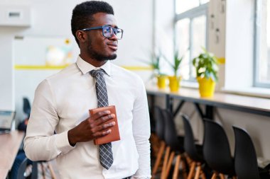 Resmi giyinmiş Afrikalı işadamı modern ofis alanında defterini tutuyor, düşünceli bir şekilde şirket başarısını ve çeşitli iş yerlerini yansıtıyor..