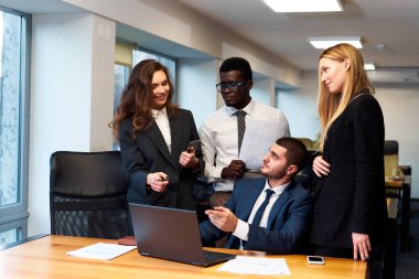 Modern ofis ortamında çok ırklı iş adamları dizüstü bilgisayarın etrafında canlı bir tartışmaya girişiyor, gülümseyen bir kadın, kağıtları olan Afrikalı bir adam, işadamı sarışın kadını dinlerken.