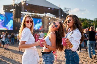 Güneş gözlüklü bir grup gülümseyen genç kadın, yaz müzik festivalinin tadını çıkarıyor. Sahilde renkli içkiler, modaya uygun günbatımı partisi havası var. Arka planda konser sahnesi ve kalabalık toplanıyor..