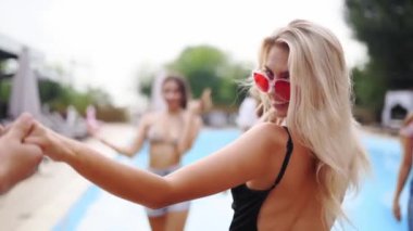 Kırmızı güneş gözlüklü sarışın kadın erkeğin elini tutuyor, gülümsüyor ve flört ediyor. Erkek, sıcak yaz gününde yüzme havuzu partisine mayolu kızları takip ederek dinlenir, yüzer ve eğlenir. Arka plan.