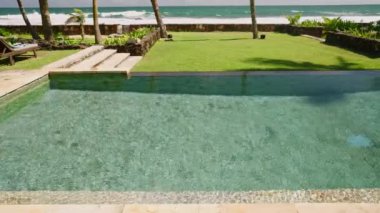 Yüzme havuzu ve tropik adada kumlu okyanus kıyısında palmiye ağaçları olan yeşil çimenli lüks bir tatil köyü. Yüzeyinde dalgalanmalar olan mavi ılık su ve turistler için rahat bir bahçe..