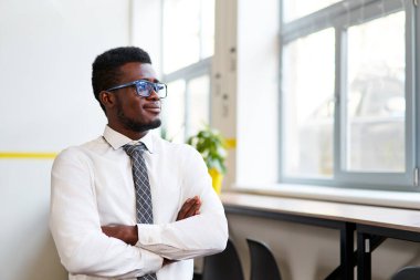 Beyaz gömlekli, kravatlı ve gözlüklü kendinden emin Afrikalı şirket çalışanı modern ofiste kollarını kavuşturarak somut liderliği ve profesyonel başarıyı temsil ediyor..