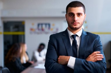 Profesyonel erkek lider, modern şirket ortamı, çok ırklı çalışanlar. Takım elbiseli kendine güvenen Arap işadamı ofiste duruyor, kolları bağlı, arka planda çalışan bir ekip..
