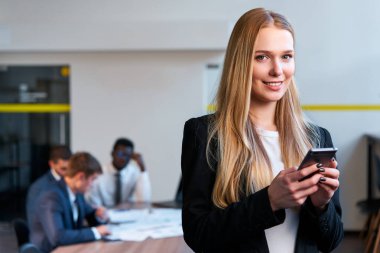 Profesyonel kadın, gündelik kıyafetlerle kurumsal görevler için mobil uygulamalarla etkileşime giriyor. Kendine güvenen sarışın iş kadını modern ofiste akıllı telefon kullanıyor, iş arkadaşları arkada çalışıyor..