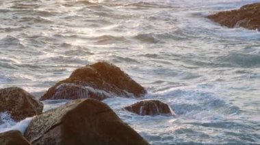 Okyanusa yansıyan altın güneş ışığıyla kıyı manzarası. Gün batımında dalgalar kayalara çarparak sahilde deniz köpüğü oluşturur. Rahatlatıcı, doğa geçmişi. Meditasyon, doğal manzara. Yavaş çekim.