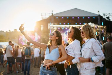 Mutlu arkadaşlar partisi, kalabalık içinde dans. Bir grup genç kadın yaz plaj müzik festivalinde selfie çekiyor. Güneş sahne arkasında batar. Kızlar konserden, tatilden hoşlanırlar. Sıradan moda, gençlik kültürü.