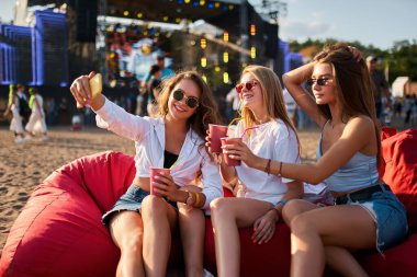 Günlük kıyafet, güneş gözlüğü, içki yudumlama, biri selfie çekiyor. Bir grup kadın plajda müzik festivalinin tadını çıkarıyor, fasulye çuvalları üzerinde uzanarak. Sıcak gün ışığı. Açık hava yaz etkinliğinin rahatlatıcı ve eğlenceli atmosferi..