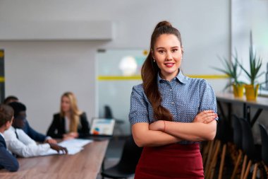 Ekip çalışması arka planda, çeşitli meslektaşlar projeyi tartışıyor. Kendine güvenen Asyalı iş kadını modern ofiste kollarını kavuşturmuş duruyor. Şirket ortamında profesyonel toplantı, liderlik, girişim.
