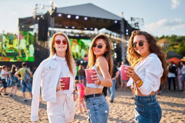 Gülümseyen kadınlar kutlama yapar, ellerinde içkilerle dans ederler. Yaz plajı müzik festivalinde bir grup kız kadeh kaldırıyor, arka planda canlı konserle güneşli bir günün tadını çıkarıyor..