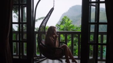Kadın sallanan sandalyede çaydan hoşlanıyor, sisli dağ tepelerine tepeden bakan manzaralı terasta dinleniyor, ve zengin yeşil bitkiler, Ella Sri Lanka 'daki lüks inzivada huzurlu bir anda temiz hava soluyor..
