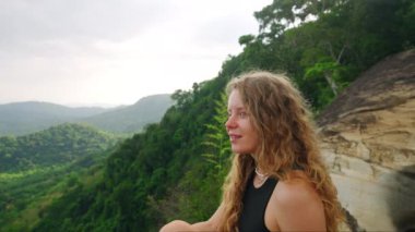 Yeşil vadi manzarasının tadını çıkarırken doğadaki dinginlik. Kadın şelalenin yamaçlarında oturuyor, uzun kıvırcık saçlar rüzgarda hafifçe savruluyor. Macera seyahati, tropikal bölgelerdeki sakin manzaraya kaçmak.