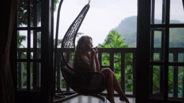 Yeşil manzaralı lüks balkon, sakin inzivaya çekilip sıcak içecek ve egzotik manzara ile canlanıyor. Kadın, dağlık çay tarlasına bakan sallanan sandalyede çayını yudumluyor..