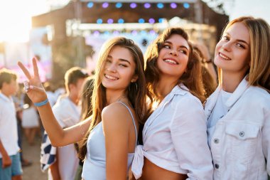 Arkadaşlar konserin tadını çıkarırlar, günbatımı ise arka planda. Bir grup mutlu kız yaz plaj müzik festivalinde, gülümsüyor, güneşin altında dans ediyor, barış işareti yapıyor. Günlük kıyafetler, eğlence, gençlik kültürü havası.