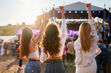 Günlük yaz kıyafetleri, günbatımı ışıkları, arka planda kalabalık ve sahne, kızlar birlikte dışarıda kutluyor. Üç genç kadının plaj müzik festivalinde içkilerinin yükselmesi ve konser havasının tadını çıkarması..
